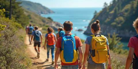 High school students hike along coastal trail on field trip, enjoying outdoor education, nature exploration. Backpacking teens walk through forest path by ocean under sunshine learning about ecology.